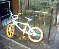 A white BMX bike with red and yellow tires leaning against a a fence